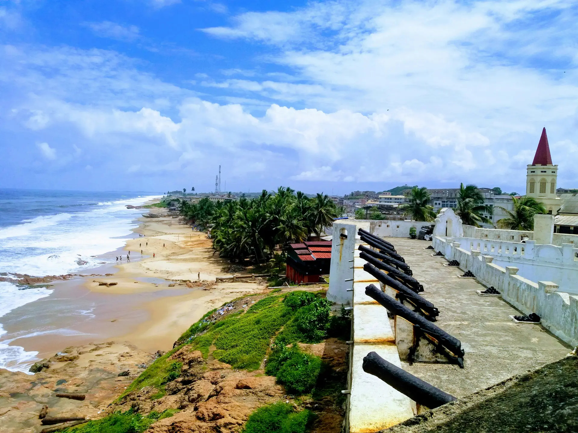 Cape Coast beach landscape featuring golden sandy shores meeting turquoise Atlantic Ocean waters, with historic coastal architecture visible along the shoreline under a bright daytime sky, capturing the serene and welcoming atmosphere of Ghana's famous coastal destination