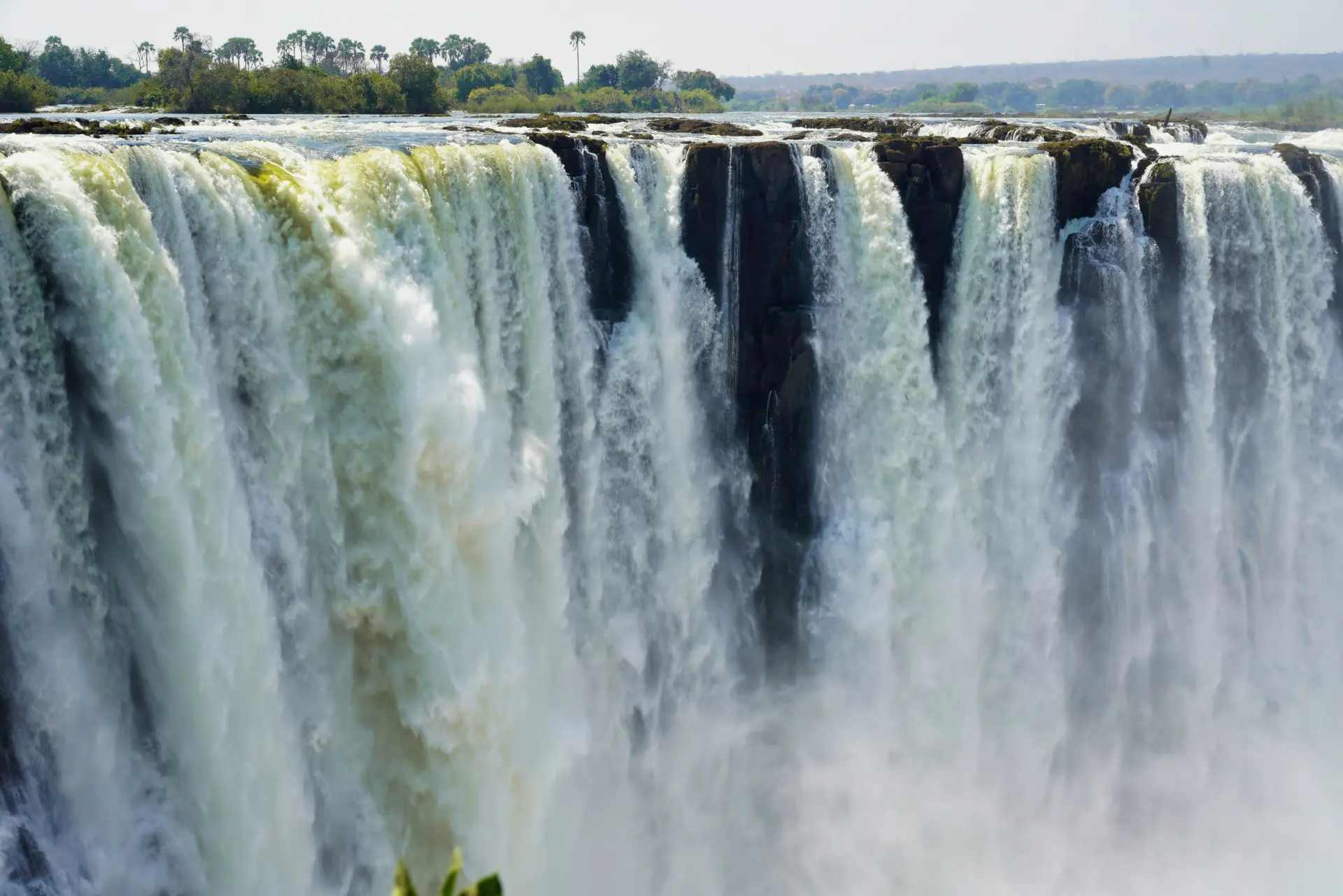 Massive Victoria Falls cascading down rocky cliffs with white mist rising from the base, surrounded by lush green vegetation under a bright sky, showcasing the powerful natural wonder straddling Zimbabwe and Zambia in Africa